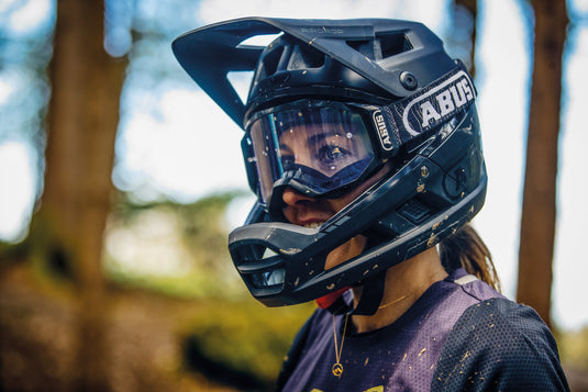 Female cyslist stading in woods wearig black ABUS AirDrop helmet and ABUS Buteo goggles