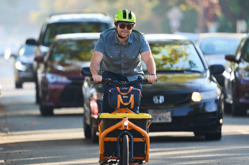 Load image into Gallery viewer, cyclist riding cargo bike down busy street wearing signal yellow ABUS HUD-Y helmet. #color_signal yellow

